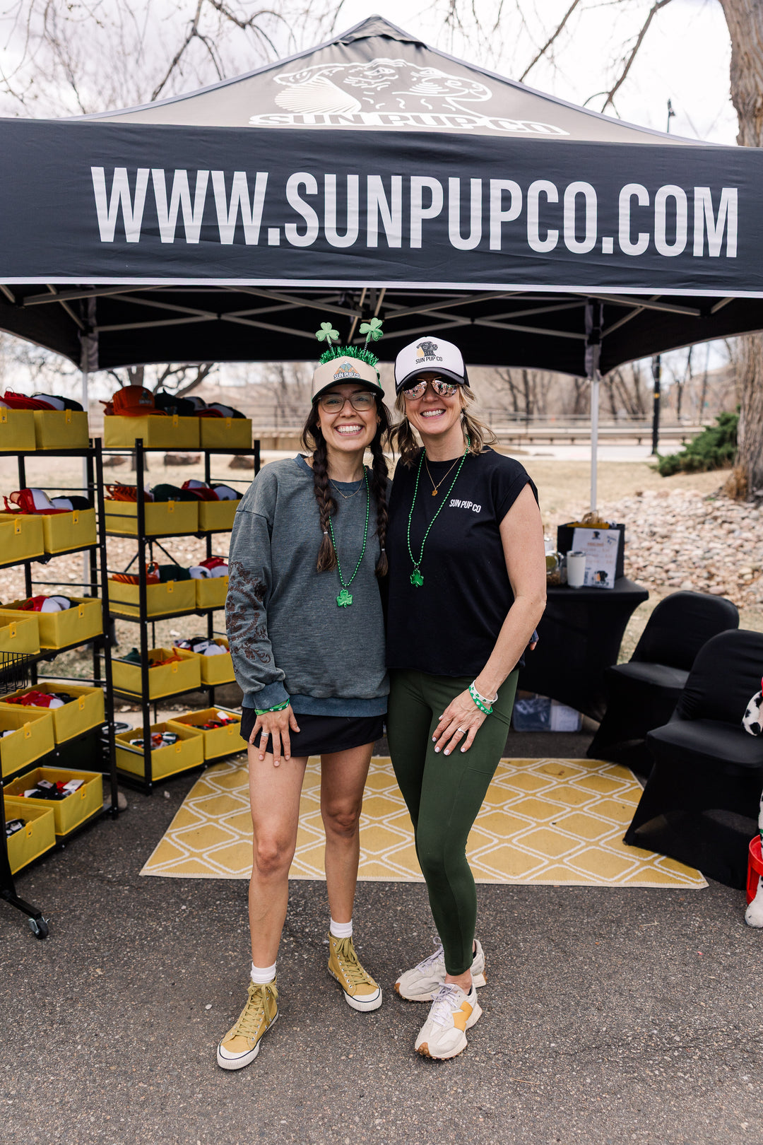 Two women standing under a tent with 'www.sunpupco.com' on it, with a store setup in the background.