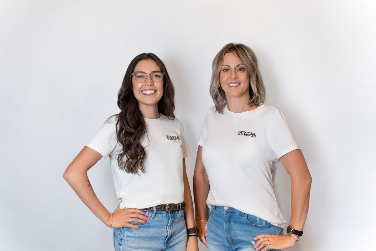 Two women standing side-by-side wearing matching white t-shirts with a black logo on the left chest and blue jeans, posing against a plain white wall.